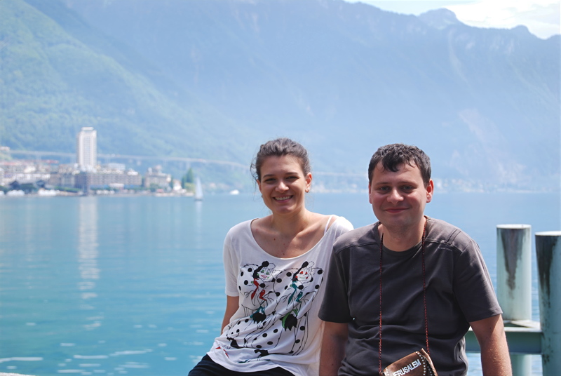 29 - Two individuals posing outdoors near a scenic alpine lake with mountains in the background, resembling the picturesque shores of Lake Geneva